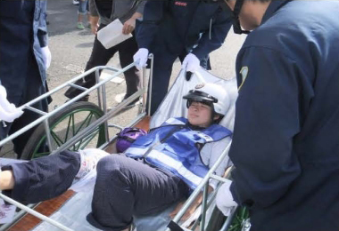 自主防災訓練の様子11。救出班により、身近にある雑誌で右足の骨折箇所を固定処置されている様子。また、警察官の気遣いによりヘルメットで頭部を保護されている負傷者。