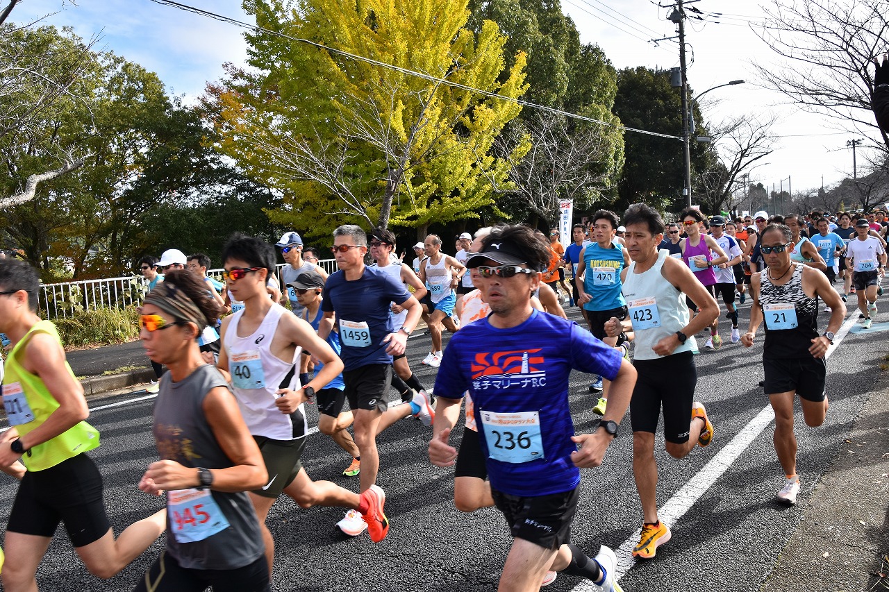 2024成田POPラン大会の10キロメートルの部に参加したランナーがスタート直後に勢いよく走る様子の写真。背景には、黄色く色付いたイチョウの木が写っている。