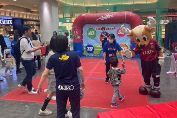 NARITAスポーツツーリズムフェス！でキックターゲットブースを設置している写真。奥にはサッカーゴールの形をした的が立っていて、手前では子どもとマスコットキャラクター「しかお」がハイタッチしている様子。