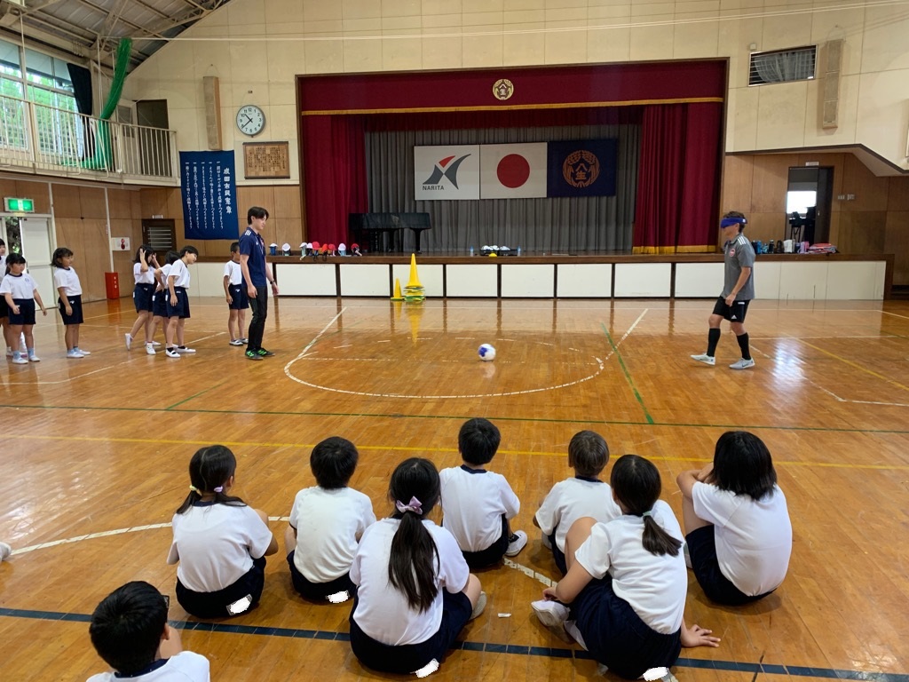 アイマスクを着けた講師が、集まった児童の前でブラインドサッカーボールを使ってパスの手本を見せる様子