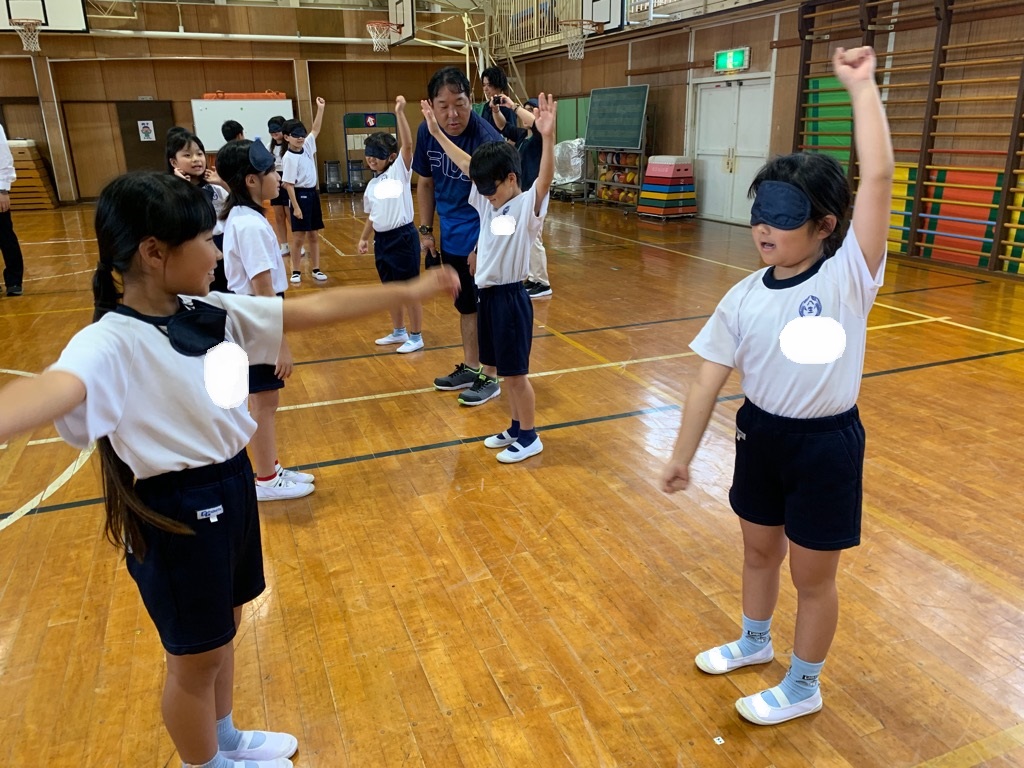 児童がペアを組み、アイマスクを着けた相手へ言葉でストレッチの動きを伝える様子