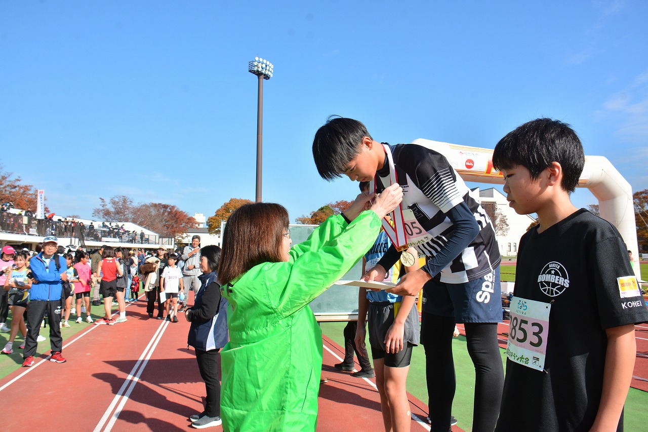 2025成田POPラン大会の表彰式での様子の写真。教育長が1位のランナーにメダルをかけている。
