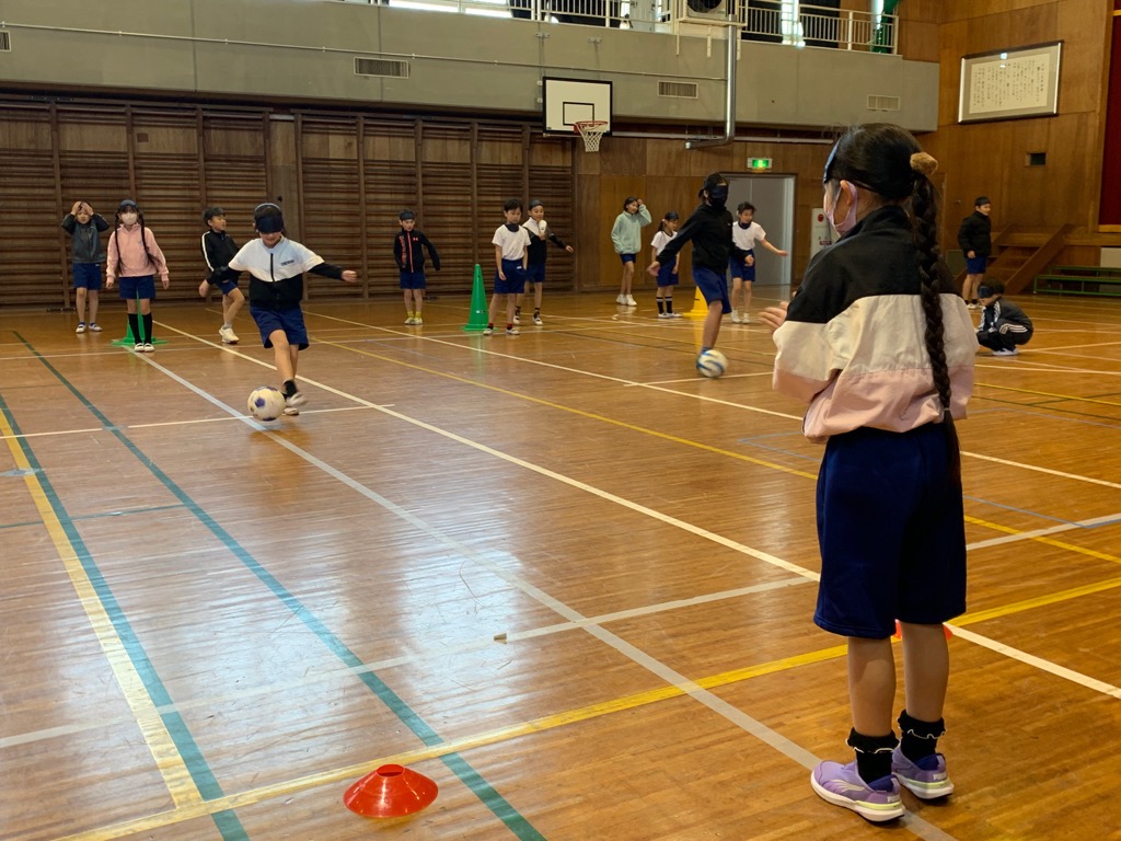 アイマスクを着けた児童が足元にあるブラインドサッカーボールを操り、仲間が拍手と呼びかけで音を出して案内する方向へパスを出す様子
