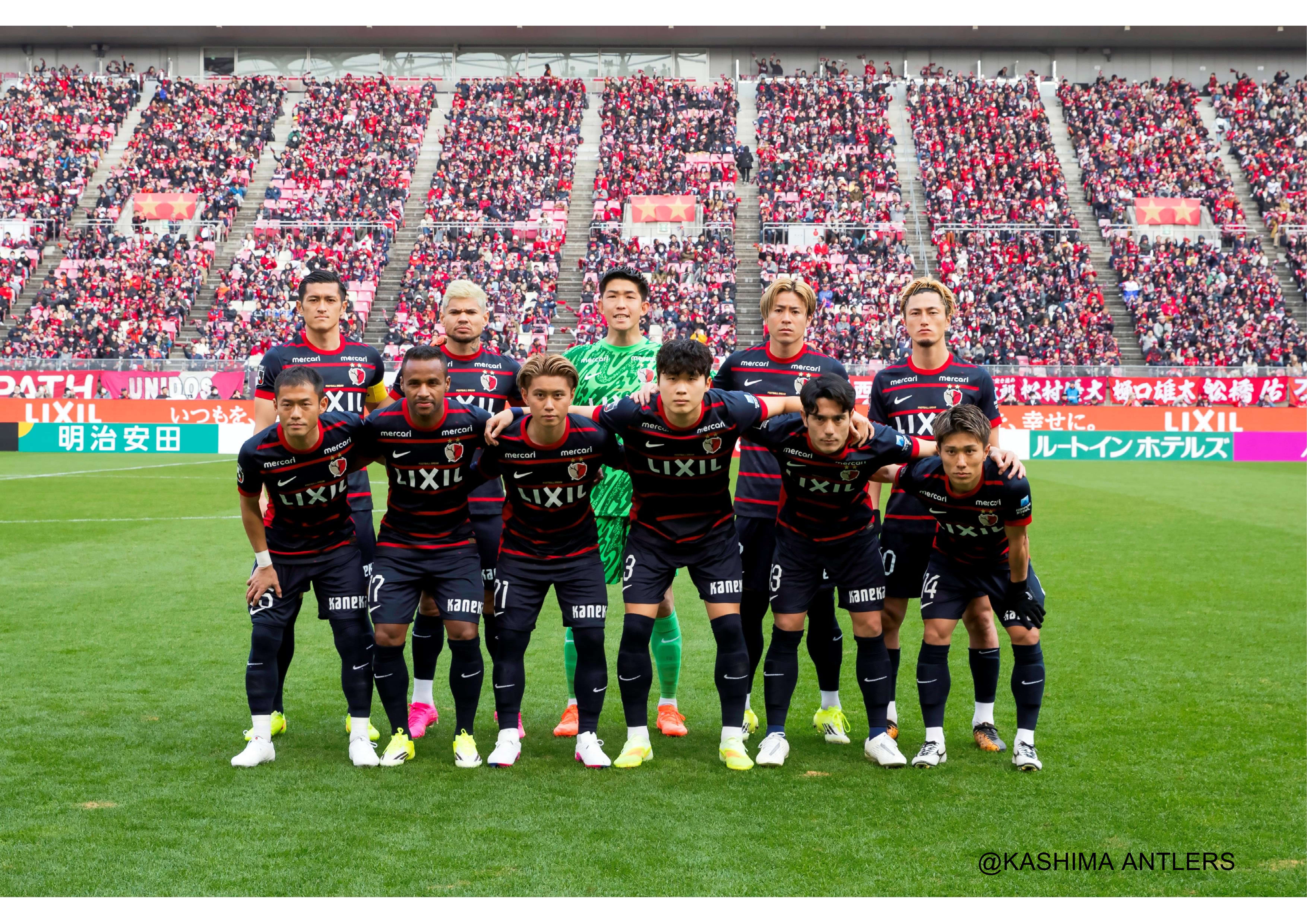 明治安田J1百年構想リーグの試合に臨む鹿島アントラーズのスターティングメンバ―を撮影した集合写真。背景の観客席には鹿島アントラーズのサポーターもたくさん写っている。