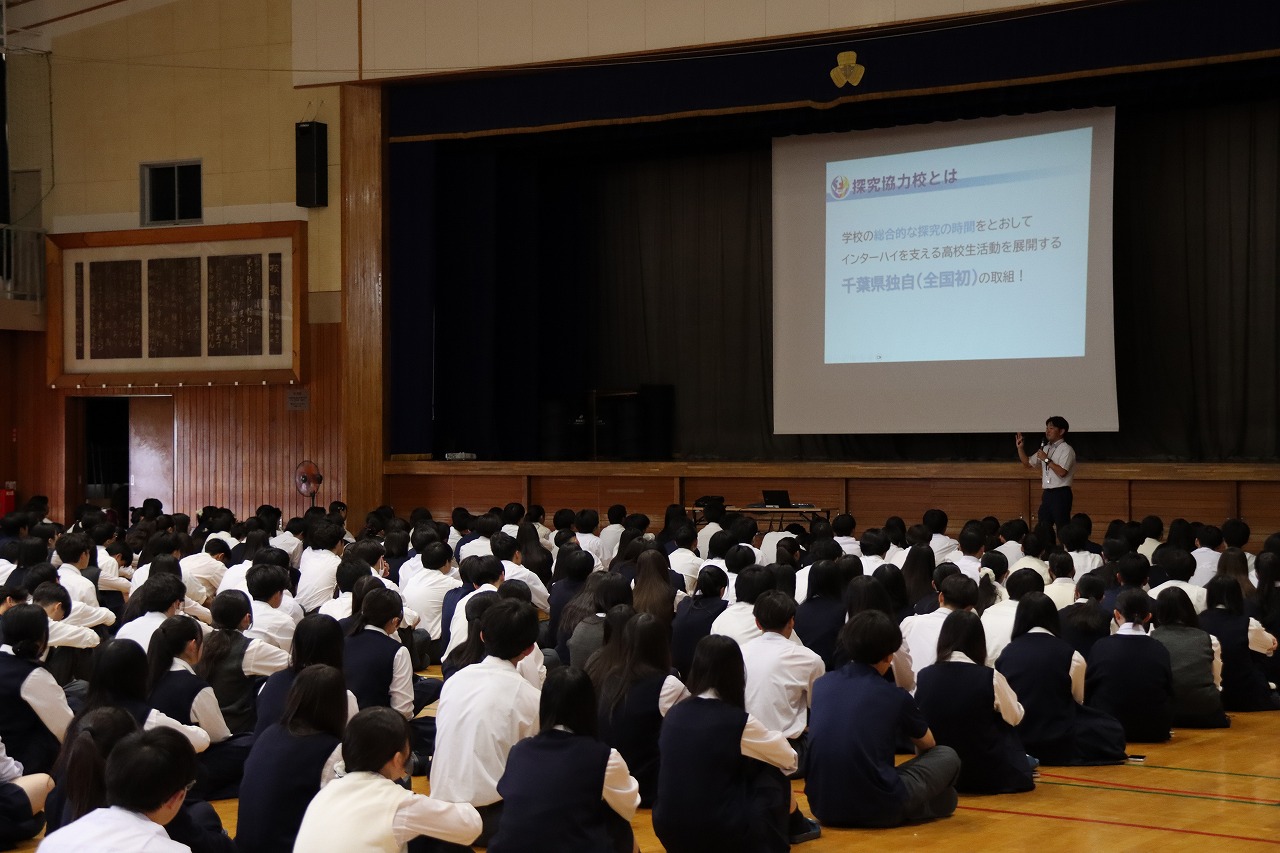 千葉県教育委員会高校総体推進室の職員が千葉県立成田北高等学校の全校生徒の前で説明を行う様子。会場の体育館で、床に座る生徒たちの前へ職員が立ち、スクリーンに映された資料を基に説明している写真。