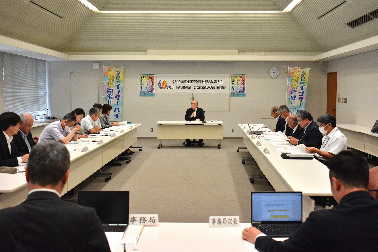 成田市実行委員会設立総会及び第1回総会の会議中の写真。正面に議長を務める小泉市長が座って議事を進行し、その前には向き合って委員が座っている。